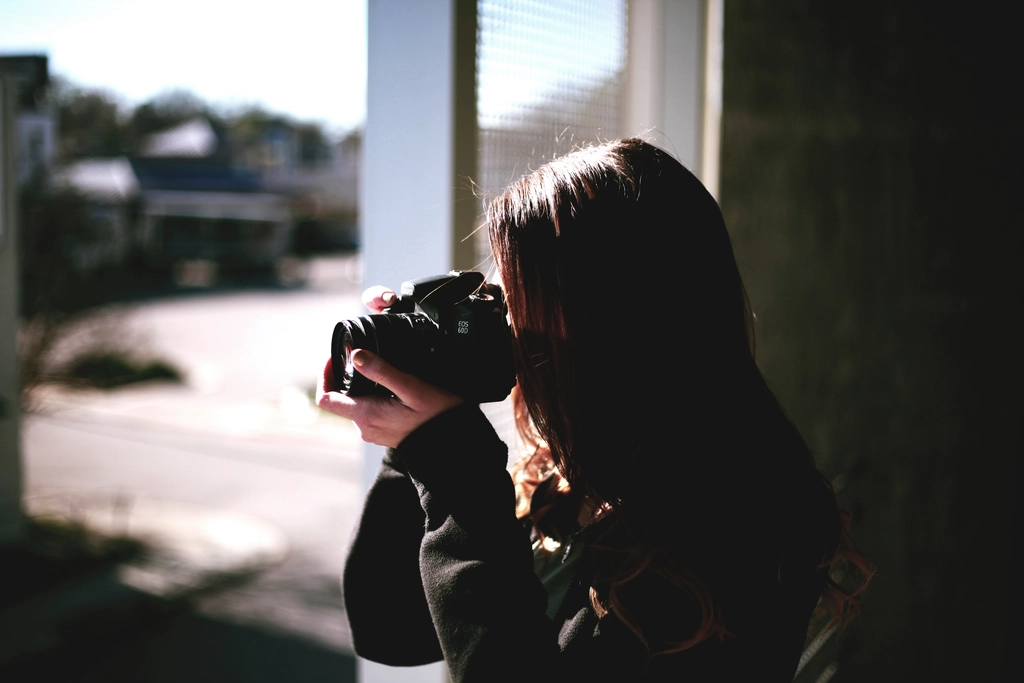 A woman taking a photo with a camera beside a sunny window